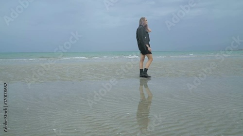 Beautiful woman walks along the beach, cold rainy weather, autumn. Sexy girl hipster or punk in a black leather jacket is walking through puddles near the sea or ocean, looks in the water reflection