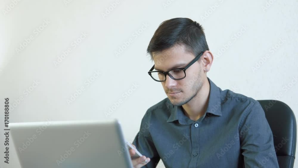 Close-up portrait of a young man wearing glasses sitting in his office in front of a monitor - working on a computer and talking on the phone. People stock footage shot. Close-up portrait of a young man wearing glasses sitting in his office in front of a monitor - working on a computer and talking on the phone. People stock footage shot.
