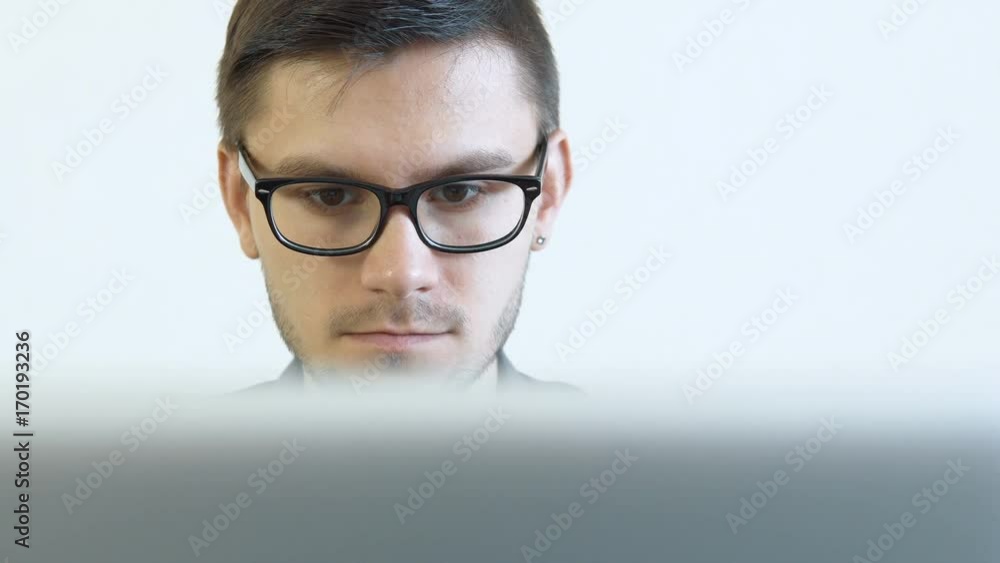 Close-up portrait of a young man wearing glasses sitting in his office in front of a monitor - working on a computer. People stock footage slider shot. Close-up portrait of a young man wearing glasses sitting in his office in front of a monitor - working on a computer. People stock footage slider shot.