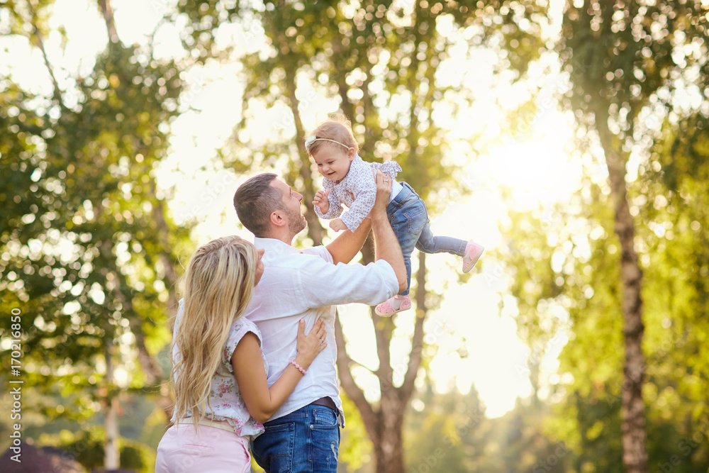 Fototapeta premium Mother father play with the baby in the park in the autumn summer. Parents raised the baby over his head.
