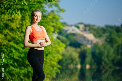 young blond woman doing exercises in nature. A sports girl in a red vest is standing in a park near the river