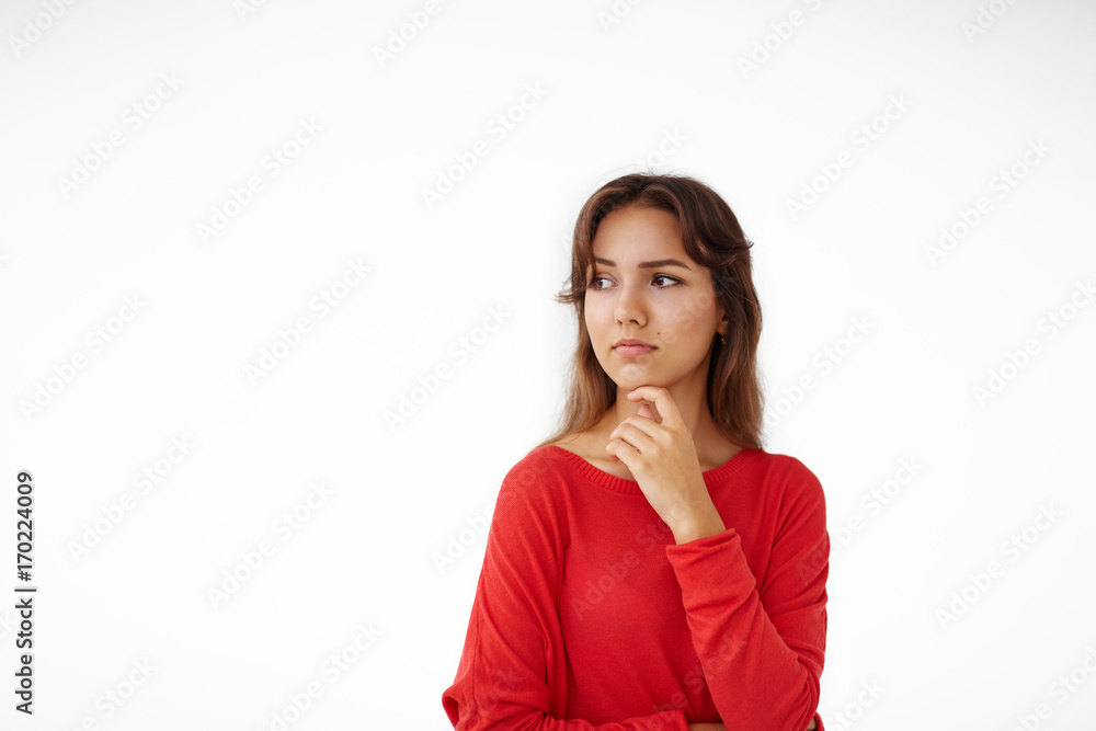 Portrait of beautiful Latin girl having thoughtful pensive look while thinking something over, looking sideways and touching he chin, posing isolated at studio wall with copy space for your content