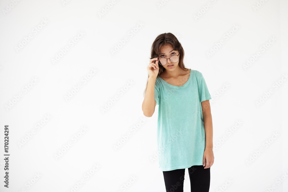 Isolated studio shot of perplexed beautiful young Latin woman with long loose natural hair wearing blue t-shirt and stylish eyeglasses peering at camera, posing at white blank copyspace wall Isolated studio shot of perplexed beautiful young Latin woman with long loose natural hair wearing blue t-shirt and stylish eyeglasses peering at camera, posing at white blank copyspace wall