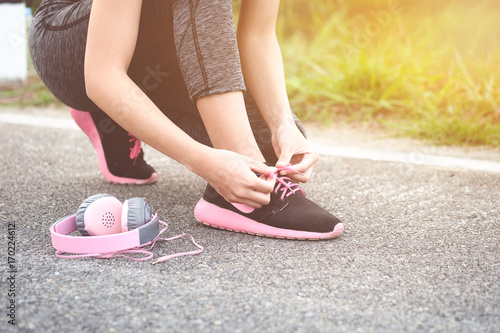 Girl runner tying laces for jogging her shoes on road in a park. Running shoes with pink headset, urban lifestyle concept