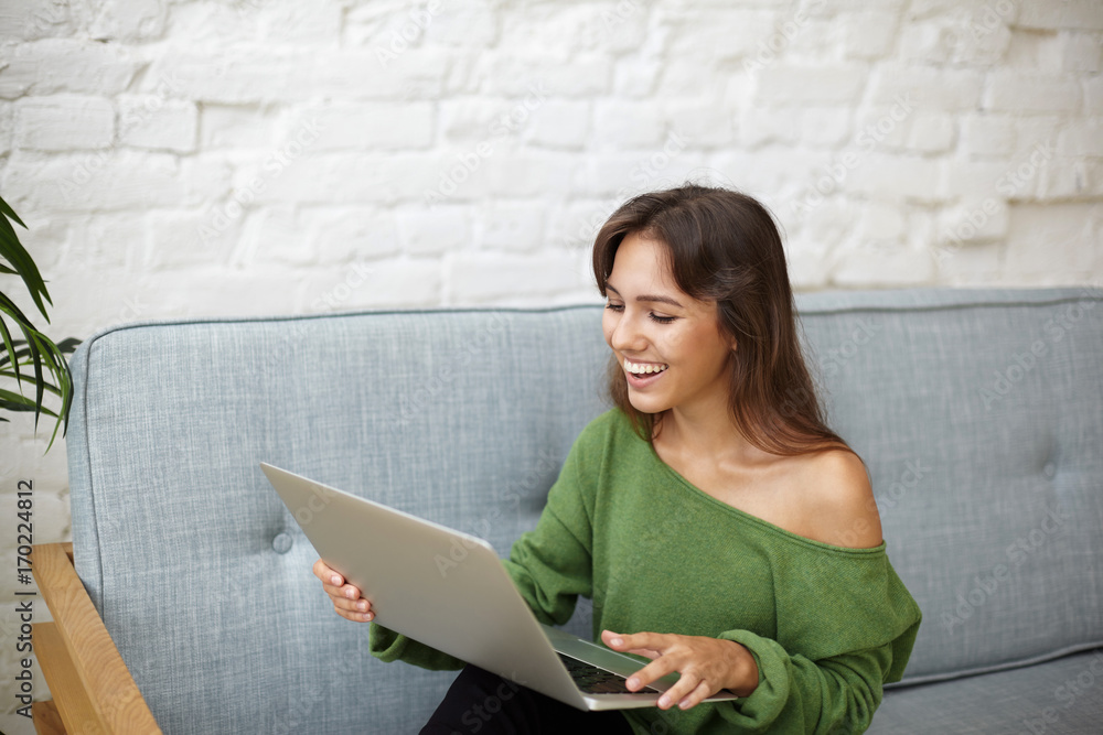 Attractive young Latin woman dressed in casual wear enjoying online communication, sitting on grey couch at home and having video conference call with friends using web enabled laptop computer Attractive young Latin woman dressed in casual wear enjoying online communication, sitting on grey couch at home and having video conference call with friends using web enabled laptop computer