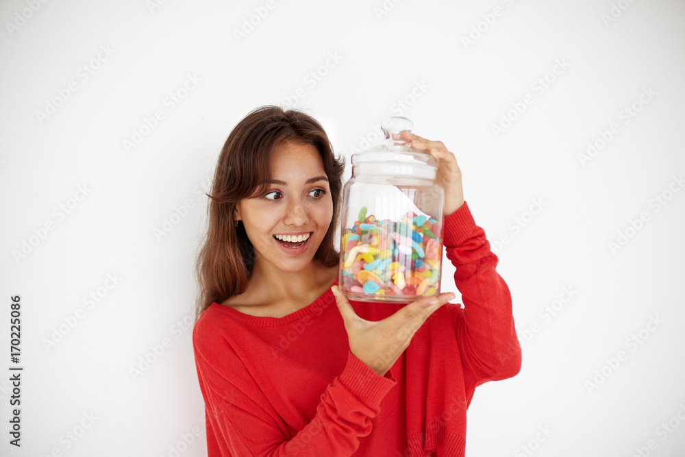 People, nutrition and unhealthy food concept. Studio shot of happy brunette mixed race girl in red top holding transparent plastic jar with colorful jelly beans, having excited and impatient look People, nutrition and unhealthy food concept. Studio shot of happy brunette mixed race girl in red top holding transparent plastic jar with colorful jelly beans, having excited and impatient look
