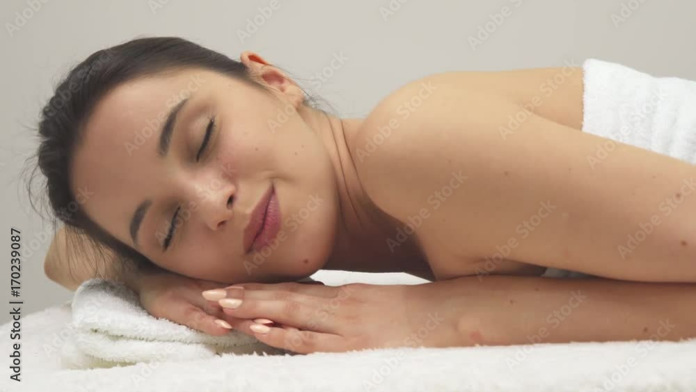 Cute caucasian girl smiling for the camera on the massage table. Attractive brunette woman falling asleep at the spa. Side view of pretty young female model closing her eyes Cute caucasian girl smiling for the camera on the massage table. Attractive brunette woman falling asleep at the spa. Side view of pretty young female model closing her eyes