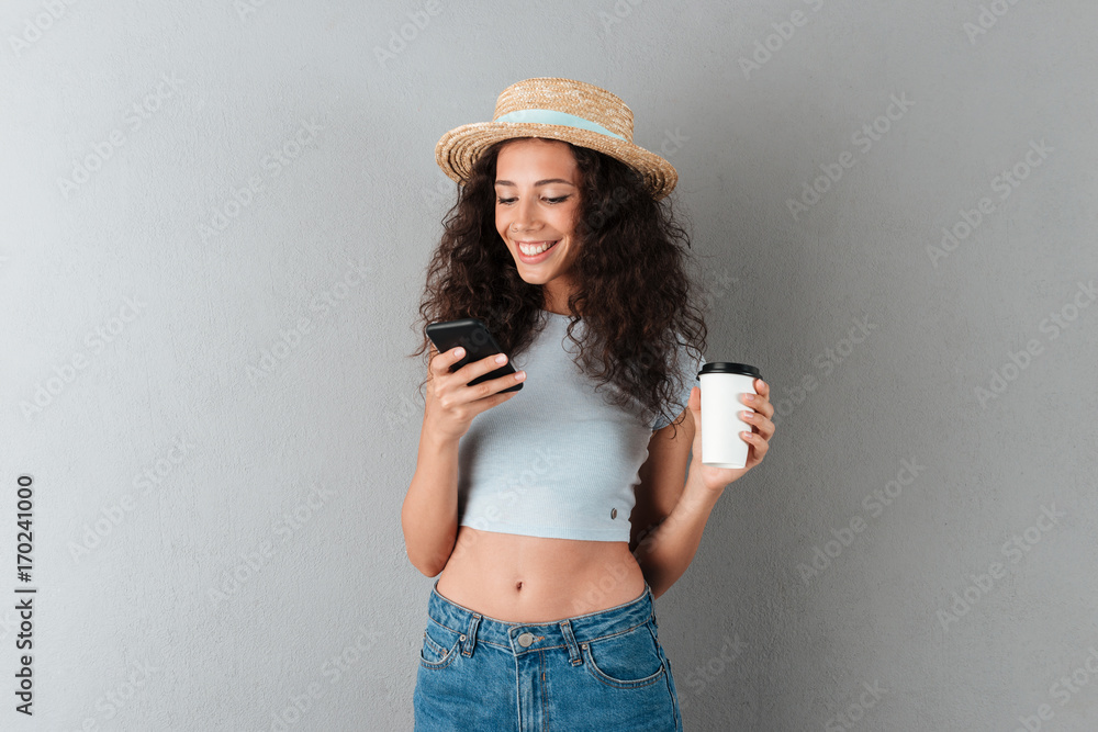 Smiling curly woman in hat using smartphone and drinking coffee Smiling curly woman in hat using smartphone and drinking coffee