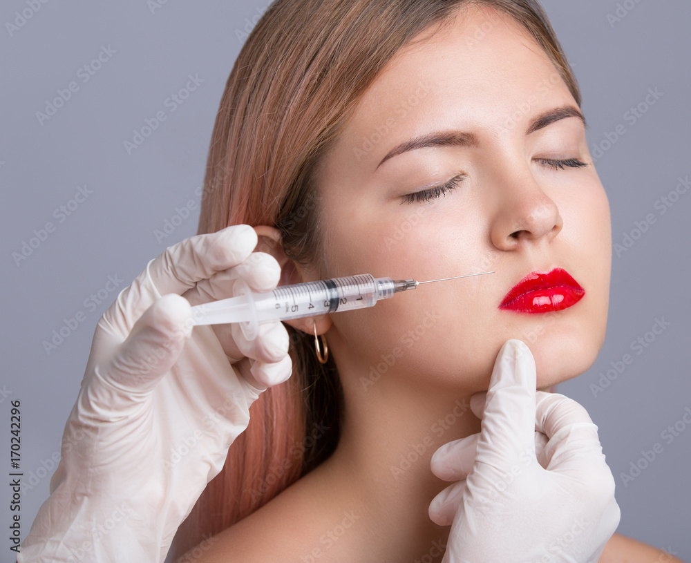 Beautiful young woman with red lips gets cosmetic injection, isolated over grey background. Doctor is making an injection in face. Beauty Treatment. Beautiful young woman with red lips gets cosmetic injection, isolated over grey background. Doctor is making an injection in face. Beauty Treatment.