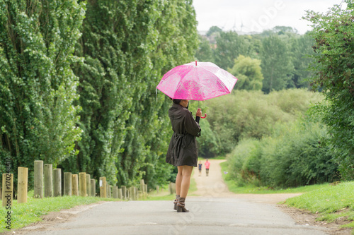 Filipino model under a umbrella walking on a pathway in a park at summertime in the rain, wearing denim shorts a black top with a coat on under a pink umbrella 