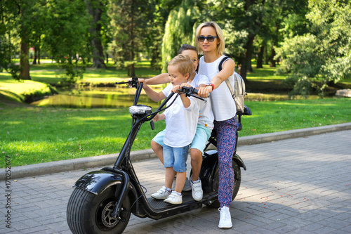 Woman with cute little boys riding electric scooter in summer park outdoors