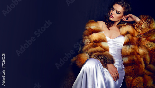 A gorgeous girl with long brown hair and a white satin dress-combination sits in a fur coat. The Snow Queen. Beauty, fashion, personal care, cosmetics, make-up, fur, fur, luxury, fur salon, luxury. photo