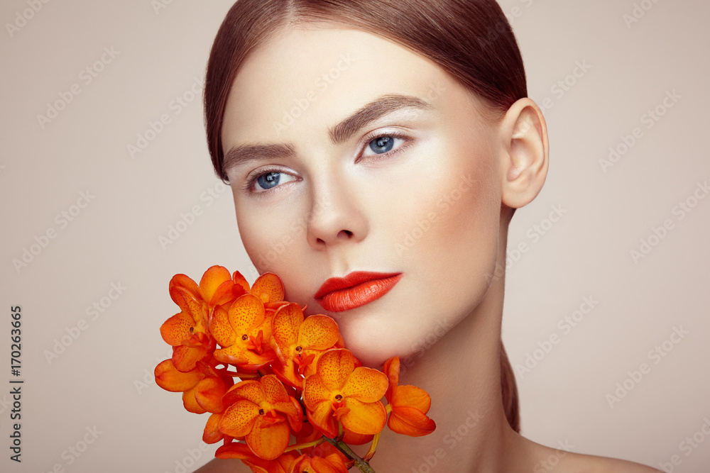 Fototapeta premium Portrait of beautiful young woman with orchid. Brunette woman with luxury makeup. Perfect skin. Eyelashes. Cosmetic eyeshadow. Orange flowers