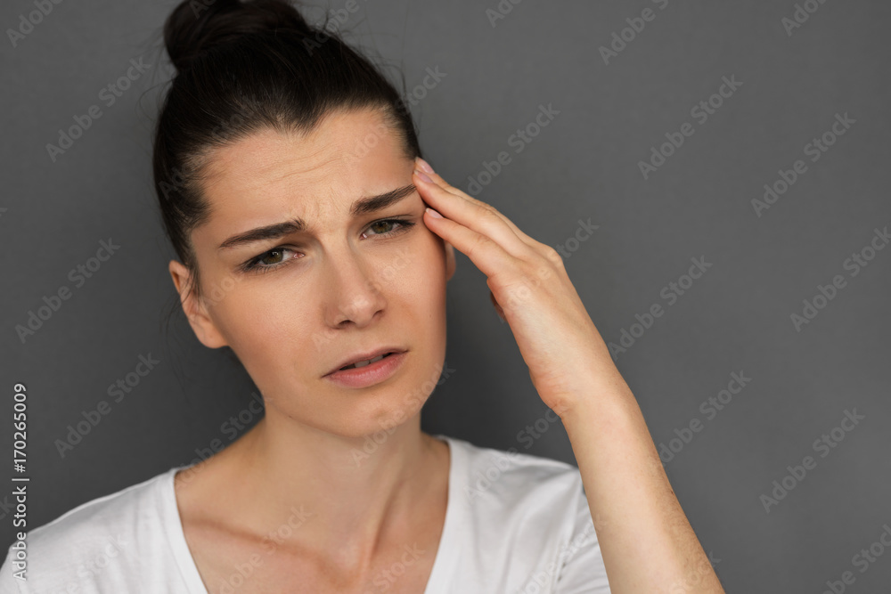 Close up headshot of unhappy young brunette female having frustrated and painful expression, touching temple with hand, suffering from bad headache or migraine while facing stress at work. Healthcare Close up headshot of unhappy young brunette female having frustrated and painful expression, touching temple with hand, suffering from bad headache or migraine while facing stress at work. Healthcare