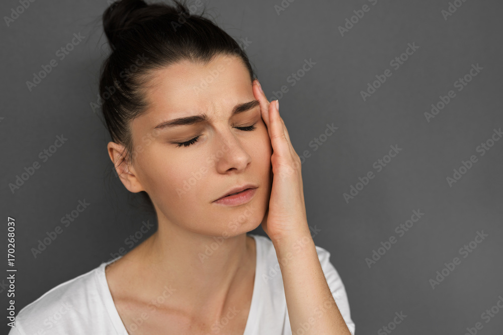 Close up portrait of sad young brunette female suffering from bad headache or migraine while facing stress at work, with closed eyes have painful expression, touching temple with hand on gray. Close up portrait of sad young brunette female suffering from bad headache or migraine while facing stress at work, with closed eyes have painful expression, touching temple with hand on gray.