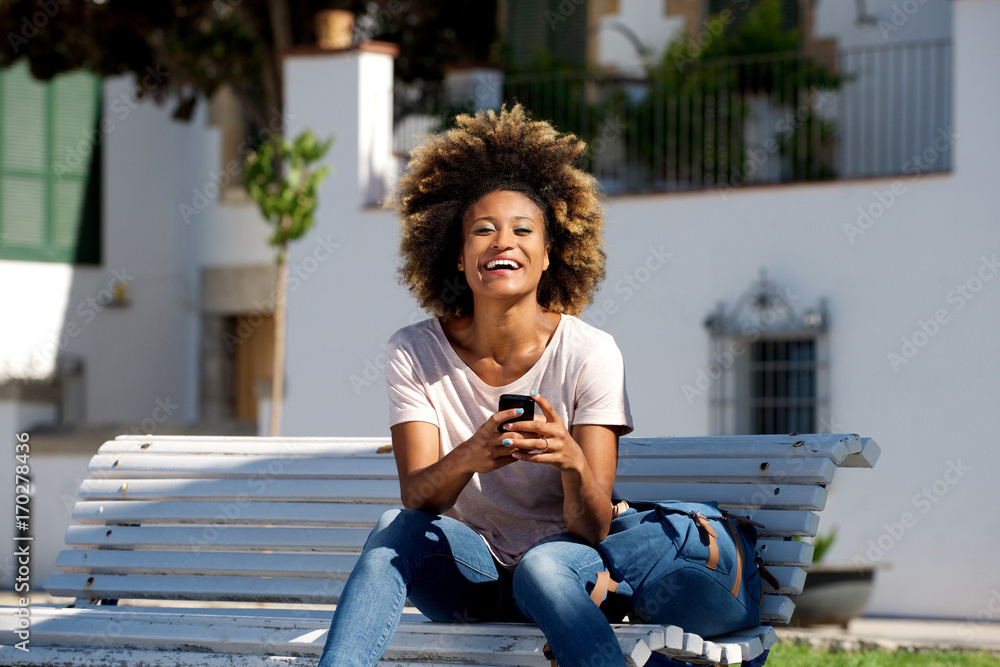 Fototapeta premium beautiful young african woman sitting outdoors on bench with mobile phone