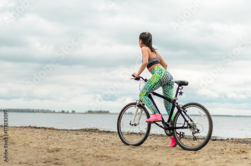 Girl beautiful brunette. Athletic body , pumped muscles. Bright colors , green ,pink . riding her bike along the shore. On the dirty sand