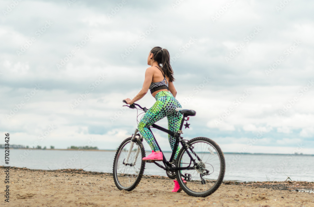 The girl the brunette plays sports on the seashore - on the beach. Rides a bike.