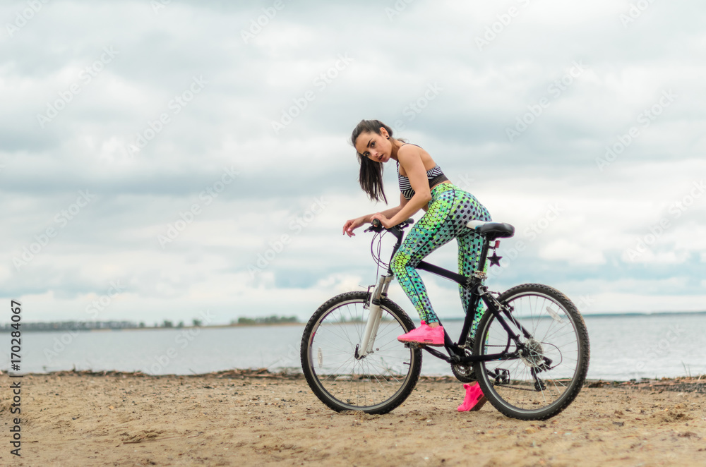 The girl the brunette plays sports on the seashore - on the beach. Rides a bike.