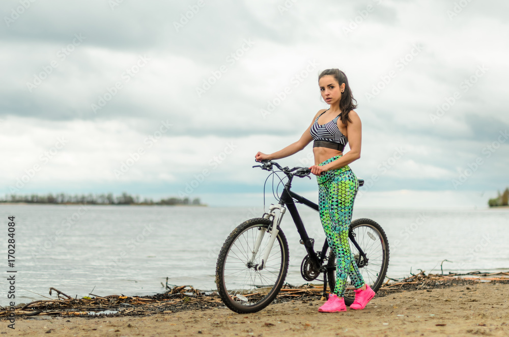 The girl the brunette plays sports on the seashore - on the beach. Rides a bike.