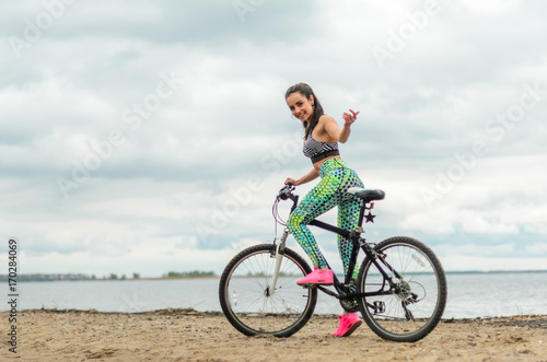 The girl the brunette plays sports on the seashore - on the beach. Rides a bike.