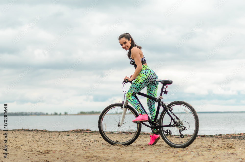 The girl the brunette plays sports on the seashore - on the beach. Rides a bike.