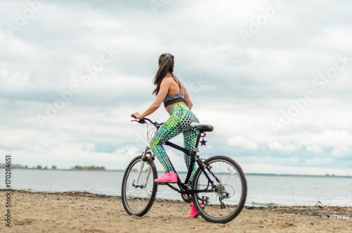 The girl the brunette plays sports on the seashore - on the beach. Rides a bike.