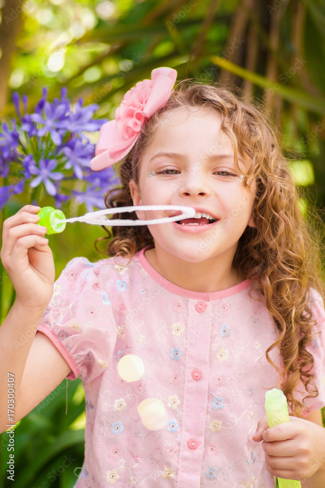 Happy little curly girl playing with soap bubbles on a summer nature, wearing a pink dress and flower in her head, in a blurred nature background Happy little curly girl playing with soap bubbles on a summer nature, wearing a pink dress and flower in her head, in a blurred nature background