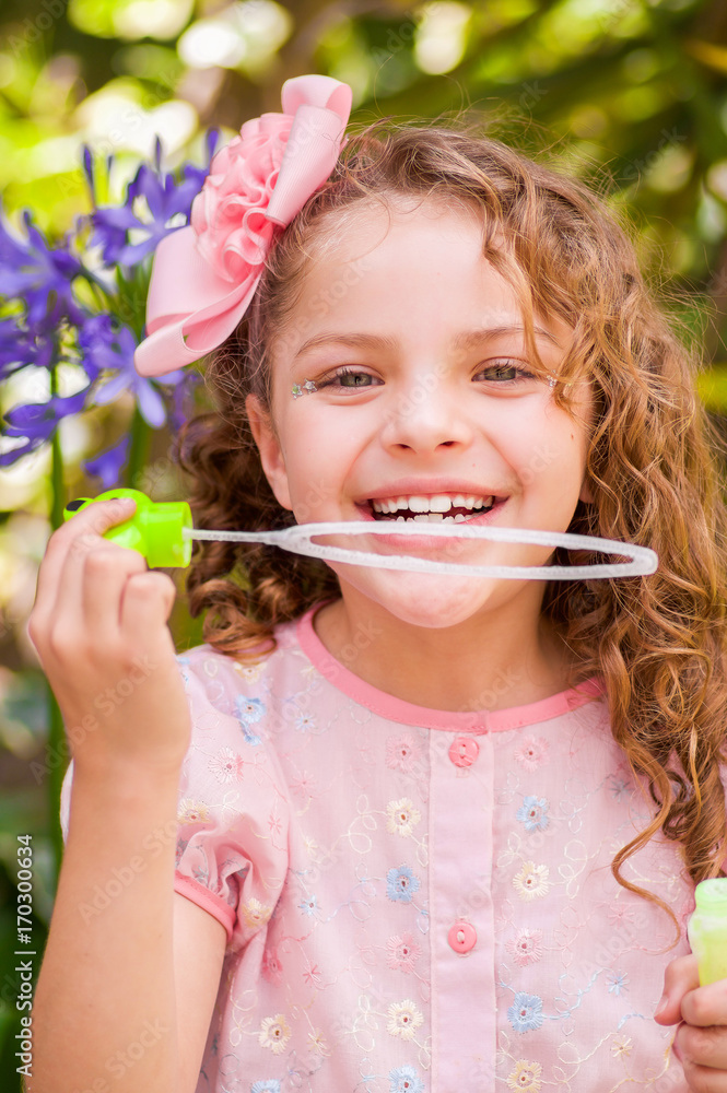 Happy little curly girl playing with soap bubbles on a summer nature, wearing a pink dress and flower in her head, in a blurred nature background Happy little curly girl playing with soap bubbles on a summer nature, wearing a pink dress and flower in her head, in a blurred nature background