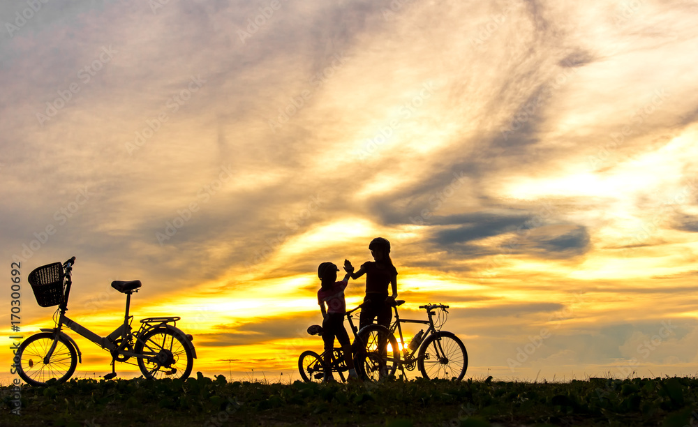 Fototapeta premium Silhouette biker lovely family at sunset over the ocean. Mom and daughter bicycling at the beach. Lifestyle Concept.