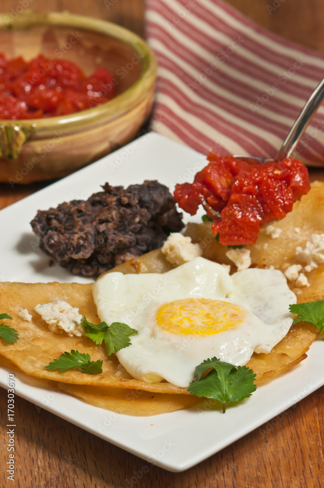 Huevos rancheros, meal, fried taco, fried egg, chopped tomatoes, refried beans, parsley on a square, white plate, bowl of tomatoes, a red and white stripped napkin, wood serving board Huevos rancheros, meal, fried taco, fried egg, chopped tomatoes, refried beans, parsley on a square, white plate, bowl of tomatoes, a red and white stripped napkin, wood serving board
