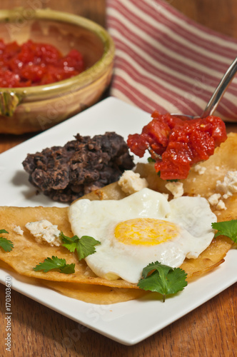 Huevos rancheros, meal, fried taco, fried egg, chopped tomatoes, refried beans, parsley on a square, white plate, bowl of tomatoes, a red and white stripped napkin, wood serving board