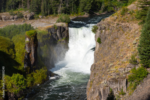 Lower Mesa Falls in the Caribou-targhee National Forest with mist being splashed onto the green foliage and rock tower.