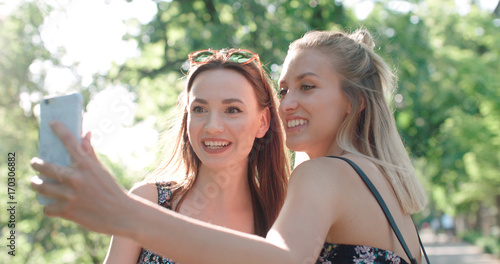 Close up portrait of two young cheerful girls having fun and making selfie, outdoors. Two beautiful teenage girls using phone in a city park. 