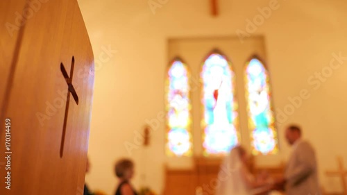 Defocsed bride and groom at wedding ceremony with christian cross in foreground on church pew photo