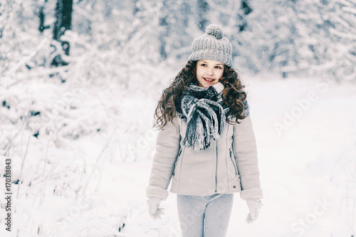 Little girl in a gray jacket with a knitted cap and scarf walking through the winter forest. Children play outdoors in a snow-covered forest.