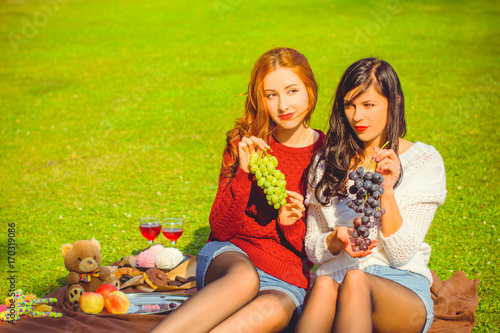 Two young pretty women with red and dark hair relax on picnic and enjoy life. Women's friendship as it is. Sisters rest on nature on the green lawn. Inspire lady with good mood at beautiful autumn day