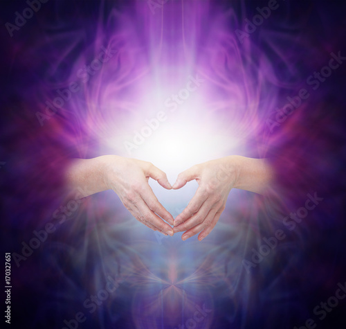 Sacred Healing Energy - female hands  emerging from a deep purple background making a heart shape over a bright glowing energy manifestation with plenty of copy space  