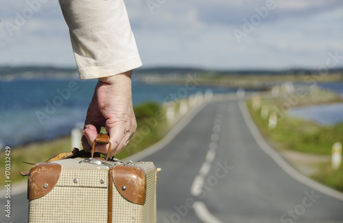 Cuadro en lienzo Senior Woman travelling on the road with her suitcase through the beautiful nature landscape with water at both sides