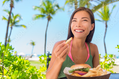 Woman eating french fries food at resort restaurant. Asian girl enjoying summer vacation at beach bar with fast food lunch meal, burger and fries.