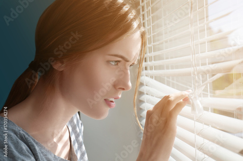 Beautiful young girl separating slats of blinds and looking through window