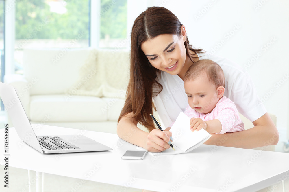 Young mother holding baby while writing in notebook in home office Young mother holding baby while writing in notebook in home office
