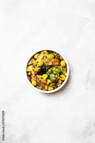 Stewed eggplants with vegetables and olives in a bowl, light background, top view