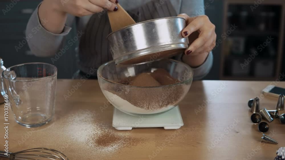 Close-up view of young woman weighs the cocoa, adds to flour. Girl using kitchen scales, bowl and sieve for this. Close-up view of young woman weighs the cocoa, adds to flour. Girl using kitchen scales, bowl and sieve for this.