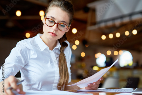 Concentrated young white collar worker wrapped up in paperwork while sitting at spacious coffeehouse, waist-up portrait shot