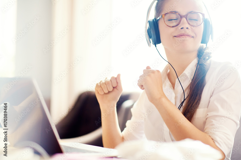 Cheerful young woman in headphones dancing to music while sitting in front of computer, blurred background Cheerful young woman in headphones dancing to music while sitting in front of computer, blurred background
