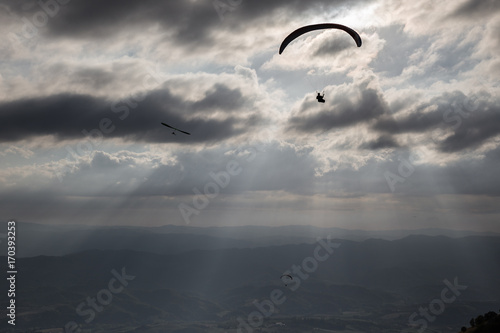Some para-gliders and hang-gliders flying over a valley, with distant mountains and hills and sun rays coming out through the clouds