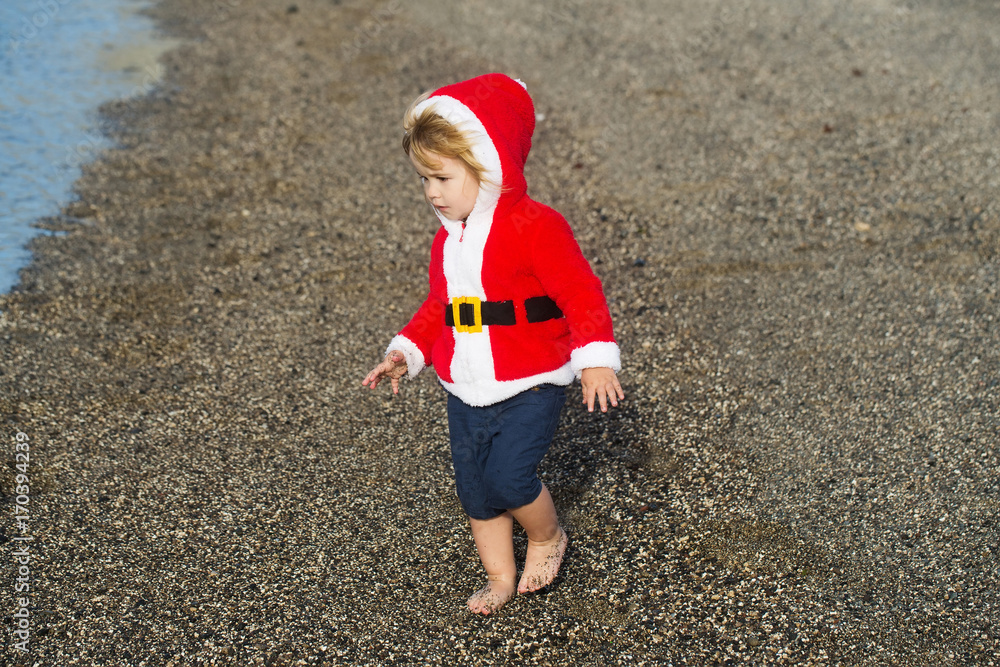 Obraz premium Cute baby boy in red santa coat on pebble beach