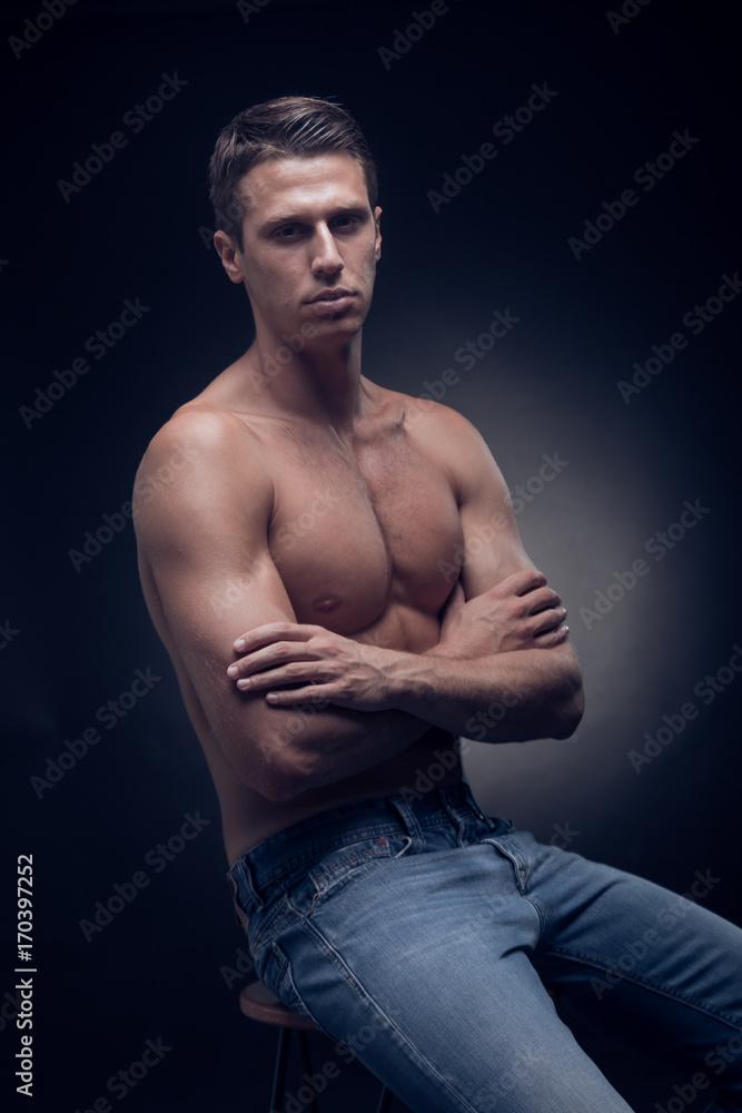 one young adult man, Caucasian, fitness model, muscular body, shirtless, jeans, black background, studio, posing, looking to camera, sitting in bar stool