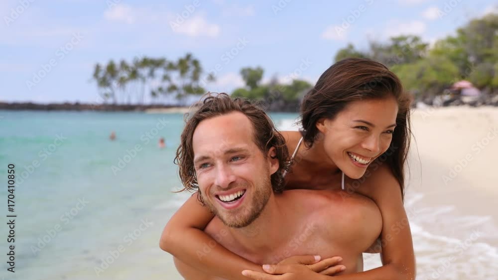Beach couple having fun. Cheerful man piggybacking woman at beach. Playful young couple is on summer vacation. Multiethnic male and female are enjoying on sunny day. Beach couple having fun. Cheerful man piggybacking woman at beach. Playful young couple is on summer vacation. Multiethnic male and female are enjoying on sunny day.